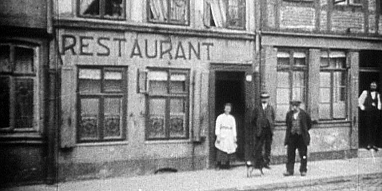 Auf dem Bild sieht man ein Restaurant in der Roten Reihe in den 1920er Jahren. Davor stehen drei Personen. Rechts in einem Hauseingang steht eine weitere Person